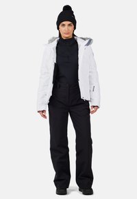 Giacca isolante bianca con accenti grigi e cappuccio foderato in pelliccia, base layer nera, pantaloni da neve neri e berretto di lana nero con pompon.