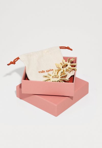 Gold hair clips in a pink box with an orange drawstring pouch. Clippings feature a sunburst design with cut-out details.