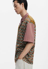Short-sleeve shirt featuring multicolored zigzag and geometric patterns, in brown, teal, yellow, and pink, with a collared neckline.