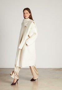Lungo cappotto di lana avorio con un colletto oversize e tasche, abbinato a pantaloni di raso beige e scarpe col tacco prune.