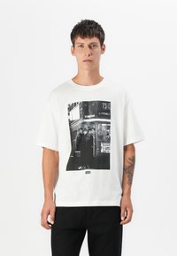 White cotton t-shirt featuring a large black-and-white graphic print of a bus and people. Short sleeves with a relaxed fit, minimalist design.