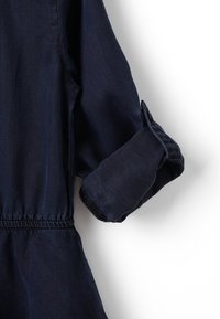 Tummansininen farkkutakki, jonka hihat on kääritty ylös, paljastaen napitettujen hihansuiden ja kokoontuneen kankaan vyötäröllä.