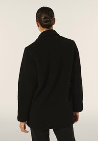 Cappotto in pile nero con un'esterno morbido e texturizzato, caratterizzato da un colletto a punte e una vestibilità rilassata. Dettagli delle cuciture visibili e maniche lunghe.