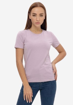 Donna con capelli castani lisci che indossa una maglietta aderente a maniche corte color lilla chiaro e jeans blu, in piedi con una mano sul fianco.