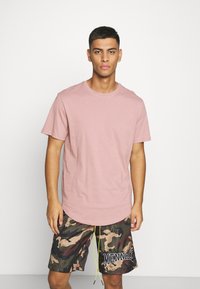 Hellrosa Baumwoll-T-Shirt mit Rundhalsausschnitt, kombiniert mit Camouflage-Shorts, die einen Kordelzug und grafischen Text am Saum haben.