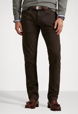 Homme portant un pantalon brun foncé, un pull gris, une ceinture marron, une montre-bracelet et des mocassins à glands marron, debout sur un fond clair.