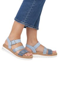 Light blue sandals with wide straps in smooth and textile materials, featuring a wedge sole and a textured cork detail around the base.