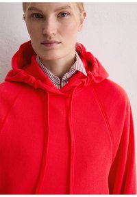 Rode hoodie gemaakt van zacht materiaal, met een trekkoord, raglanmouwen, en gedragen over een wit shirt met rode strepen.