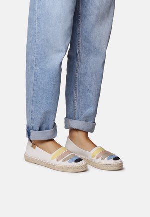 Piedi che indossano espadrillas bianche con strisce multicolori in suede abbinate a jeans azzurri risvoltati su uno sfondo bianco.