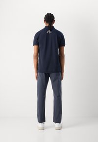 Polo shirt bleu marine à manches courtes, col classique et détail de logo dans le dos ; associé à un pantalon gris et des baskets blanches.