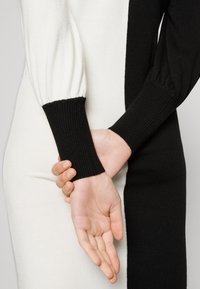 Stickad klänning med kontrasterande vita och svarta sektioner, ribbade ärmslut och ballongärmar. Slätheten på tyget med en figurnära siluett.