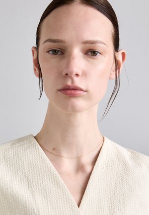 Top creme em decote em v, com um tecido texturizado; modelo usa um delicado colar de corrente dourada. Fundo neutro, iluminação suave destaca os detalhes.