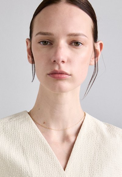 Crème v-hals top van een gestructureerde stof; model draagt een delicate gouden ketting. Neutrale achtergrond, zachte verlichting accentueert de details.