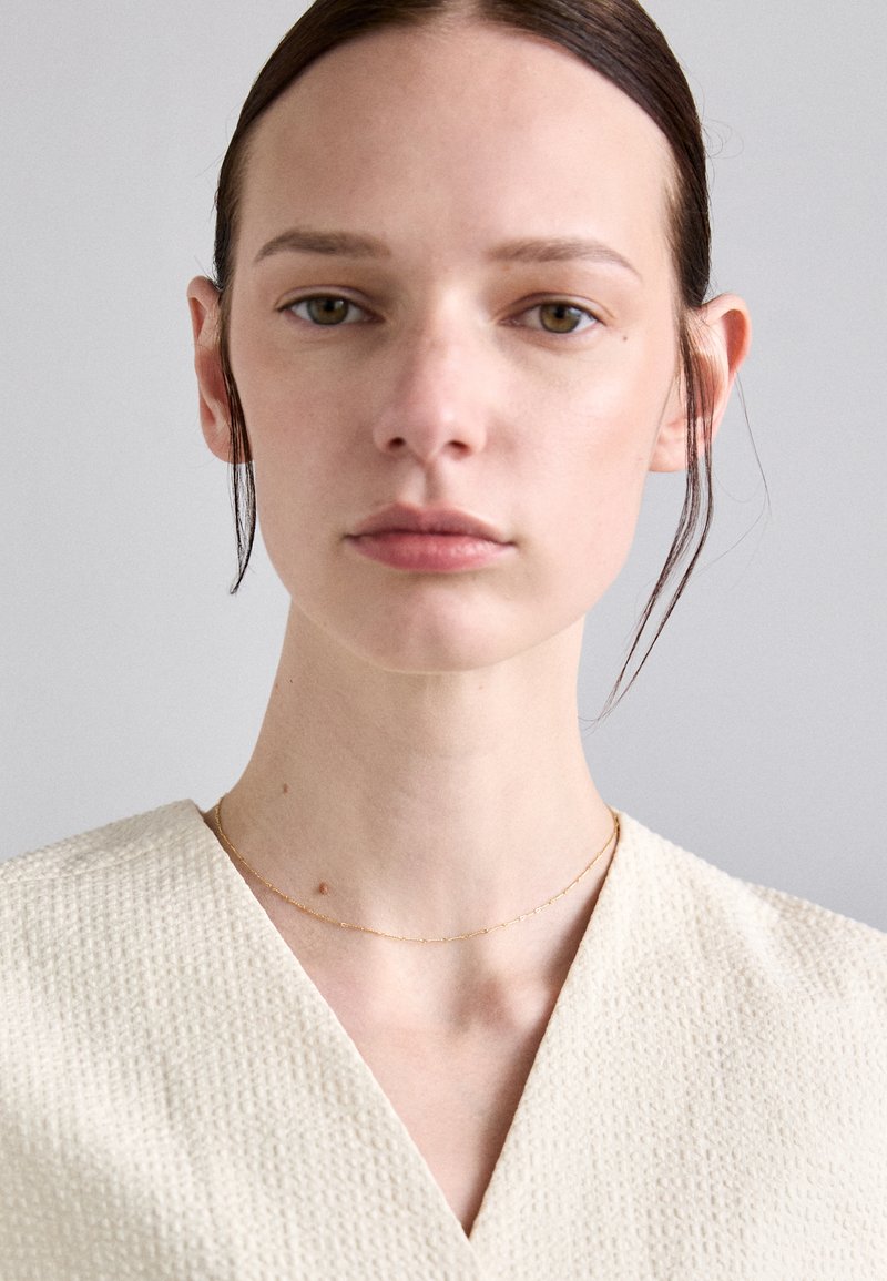 Top crem cu decolteu în V, realizat dintr-un material texturat; modelul poartă un lănțișor de aur delicat. Fundal neutru, iluminare blândă care pune în evidență detaliile.