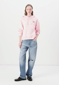 Un hoodie rose clair avec poche avant et cordon de serrage, associé à un jean bleu clair déchiré. Modèle portant des chaussures noires. Fond simple.