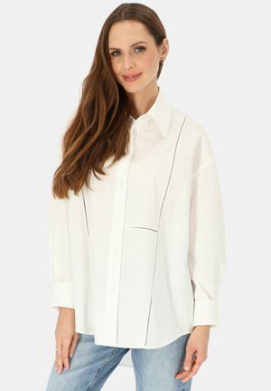 Kobieta z długimi brązowymi włosami, ubrana w luźną białą koszulę na guziki z czarnym przeszyciem i jasnoniebieskie dżinsy, stojąca na jednolitym tle.