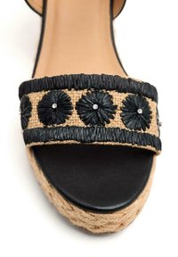 Sandalia de tiras tejidas negras con base de yute natural, con adornos florales y detalles de rhinestones, suela plana y diseño de punta abierta.