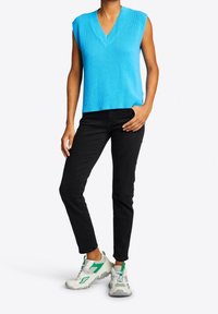 Blauer ärmelloser Pullover mit V-Ausschnitt, kombiniert mit schwarzen Slim Jeans und weißen Sneakers mit grünen Akzenten und strukturierten Sohlen.