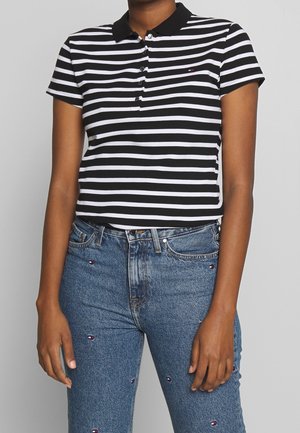 Kvinde iført en sort- og hvidstribet kortærmet polo, stoppet ned i blå jeans med små broderede logoer.