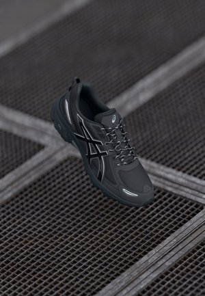 Chaussure de course noire Asics avec détails réfléchissants et lacets à motifs reposant sur une surface en grille métallique sombre.