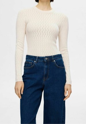 Vrouw die een witte, gebreide top met lange mouwen draagt met een zigzagpatroon en een donkere, hooggetailleerde spijkerbroek in blauw.