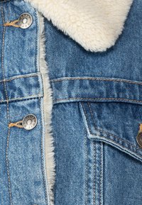 Blå denimjacka med kontrasterande vit fleecefoder och silverfärgade metallknappar, detaljerad sömnad och synlig ficka med flik.