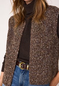 Gebreid vest in bruin met een gestructureerd, meerkleurig patroon. Heeft een open voorkant en is gecombineerd met een donkere coltrui en aangesloten spijkerbroek.