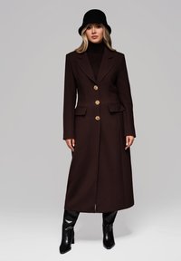 Brun uldfrakke med et langt design, fire guldfarvede knapper og to frontlommer. Kombineret med en sort bøttehat og knæhøje støvler.