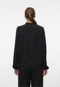 Blusa negra de manga larga con una tela texturizada, cuello con volantes y hombros abullonados. Corte holgado, combinada con pantalones negros.