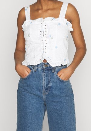 Frau trägt ein weißes, ärmelloses Oberteil mit floraler Schnürung und kombiniert es mit hoch taillierten blauen Jeans, die Hände in die Taschen gesteckt.