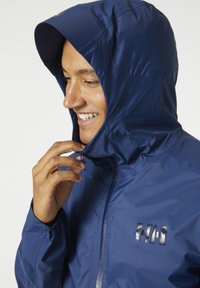 Navigatieblauwe, lichte jacket met een aansluitende capuchon, ritssluiting en logo op de borst. Glad, waterafstotend materiaal met een subtiele textuur.