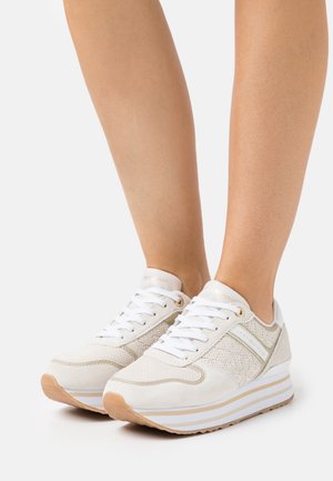Personne portant des baskets à plateforme blanches avec des accents beiges et des lacets blancs, debout sur un fond clair uni.