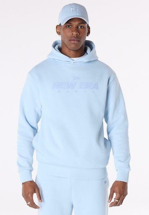 Mann trägt einen hellblauen Hoodie mit der Aufschrift "New Era", dazu passende hellblaue Jogginghosen und eine hellblaue Kappe, stehend vor einem schlichten Hintergrund.