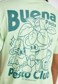 Svijetlo zelena majica s kratkim rukavima koja prikazuje crtani dizajn s velikim tekstom "Buena Pasta" i razigranim ilustracijama hrane i začina.