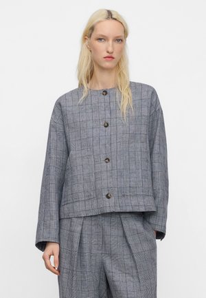 Donna bionda che indossa una giacca grigia a quadri con bottoni e pantaloni a pieghe abbinati, in piedi davanti a uno sfondo bianco semplice.