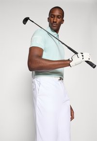 Lys myntgrønn polo-skjorte med teksturert skulderdesign, kombinert med hvite bukser. Figuren holder en golfkølle og har på seg en hvit hanske.