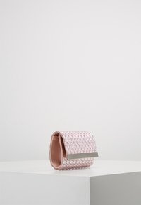 Pochette rose ornée de perles avec un accent en métal argenté reposant sur un socle blanc sur un fond gris uni.