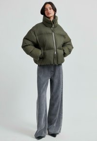 Olivgrön pufferjacka med hög krage, dragkedja och elastiska ärmslut, kombinerad med vida grå denimjeans.