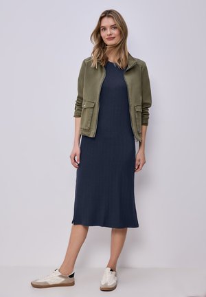 Junge Frau steht, trägt ein marineblaues Midikleid, eine olivgrüne Jacke und weiße Turnschuhe, vor einem schlichten hellen Hintergrund.