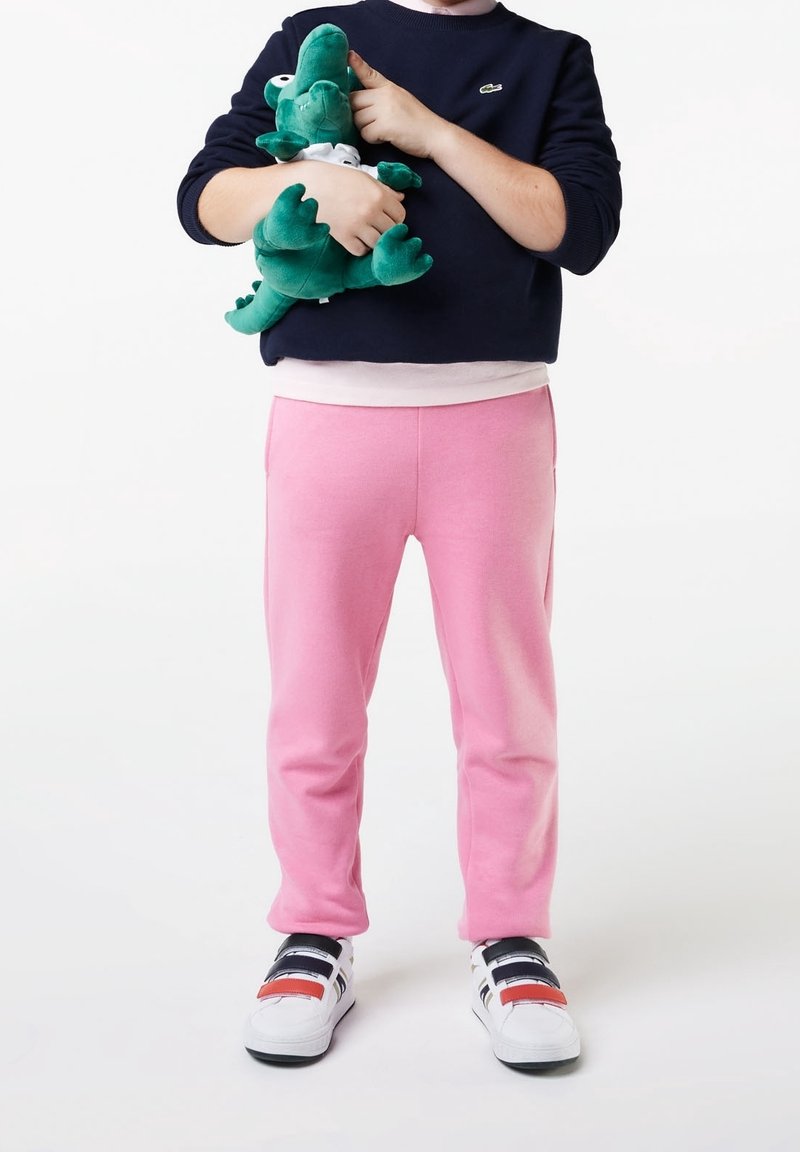 Enfant portant un pantalon rose et un pull noir tenant un crocodile en peluche vert sur fond blanc.