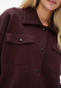 Bordowa koszula z kołnierzem, posiadająca dwie przednie kieszenie i czarne zatrzaski, wykonana z teksturowanego dzianinowego materiału.