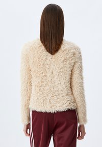 Beige Kunstpelzjacke mit einer strukturierten, flauschigen Optik, langen Ärmeln und einem schmalen Saum. Kombiniert mit burgunderroten Jogginghosen.
