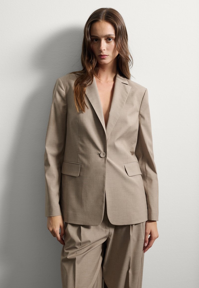 Blazer beige ajusté avec une fermeture à un bouton, revers crantés et deux poches avant, associé à un pantalon ample assorti.