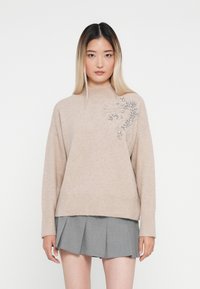 Maglione a maglia beige con collo alto, caratterizzato da un motivo decorativo argento su un lato. Abbinato a una gonna grigia plissettata.