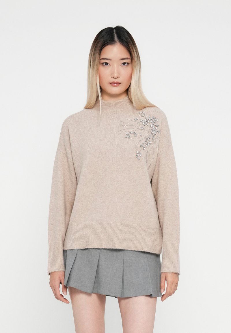 Maglione a maglia beige con collo alto, caratterizzato da un motivo decorativo argento su un lato. Abbinato a una gonna grigia plissettata.