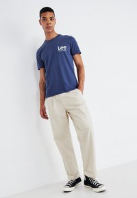 Granatowa bawełniana koszulka z małym białym logo na piersi, zestawiona z luźnymi beżowymi spodniami i czarnymi sneakersami.