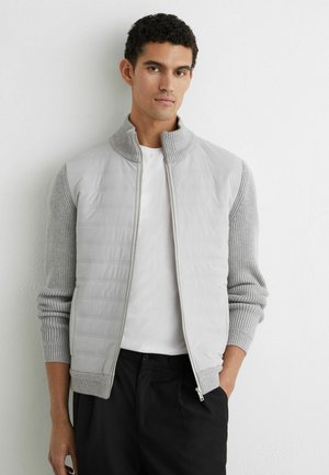 Jeune homme aux cheveux courts et bouclés portant une veste zippée gris clair sur une chemise blanche et un pantalon noir, debout contre un mur blanc.