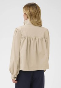 Šviesiai ruda švarka, pagaminta iš lengvos medžiagos, su aukštu apykakle, susiraukšlėtu nugaros dizainu, ilgomis pūstomis rankovėmis ir mygtukais užsegamais manžetais.