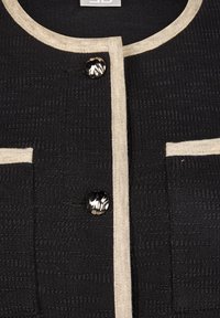 Schwarze strukturierte Jacke mit cremefarbenem Besatz, die zwei Fronttaschen und einen runden Ausschnitt hat. Knopfverschluss mit einer glänzenden schwarzen Oberfläche.