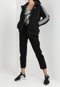 Donna che indossa una giacca Adidas nera con strisce bianche, pantaloni cropped neri, sneakers Adidas in bianco e nero e un top rosa con stampa di foglie verdi.